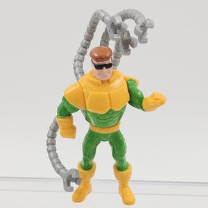 May include: A Dr. Octopus action figure. The figure is wearing a yellow and green outfit with grey mechanical arms. The figure has brown hair and sunglasses. The figure is standing on a white surface.