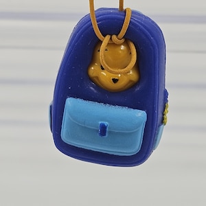 May include: A blue backpack-shaped pendant featuring a yellow bear character face peeking out from the top opening. The accessory includes a light blue front pocket with a central tab and is suspended by a thin, mustard-colored cord for hanging.