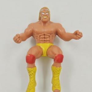 May include: A vintage action figure of a muscular wrestler. The figure is tan with yellow trunks, yellow boots, and red knee pads. The wrestler has blonde hair and a mustache, with arms outstretched.