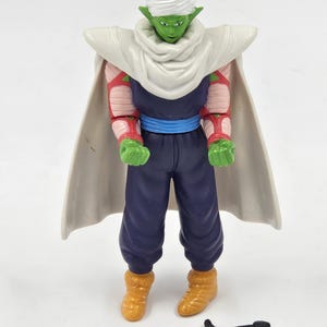 May include: A green and white action figure of a character from the Dragon Ball Z anime series. The figure is wearing a white turban, a white cape, and a blue belt. The figure is holding a sword in its right hand.