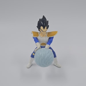 May include: A blue and yellow action figure of a character from the Dragon Ball Z anime series. The figure is holding a blue energy sphere in its hands.