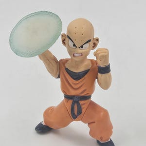 May include: A bald, orange-clad action figure with a serious expression holds a translucent, light blue disc in his right hand. The figure is posed in a fighting stance with his left fist clenched.