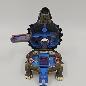 May include: A dark green turtle-shaped playset that opens to reveal a blue interior with various control panels, stickers, and a central circular opening. A small, articulated blue and purple action figure stands in front of the open toy.