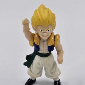 May include: A small, plastic action figure of a character with spiky yellow hair, wearing a blue and gold vest, white pants, and black shoes. The figure is posed with one arm raised in a fist.