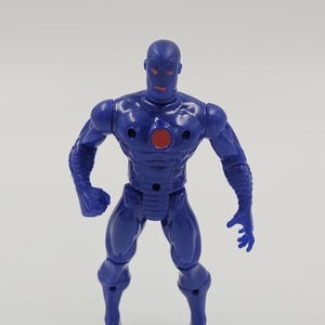 May include: A blue action figure of a humanoid robot with a red circular chest emblem and red eyes. The figure is standing with its arms slightly bent and its hands open. The figure has a smooth, glossy finish.