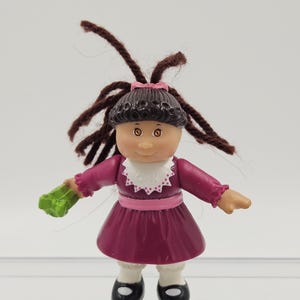 Puede incluir: Pequeña muñeca de plástico con pelo de hilo marrón, vestida con un vestido burdeos con cuello blanco y zapatos negros. La muñeca sostiene un objeto verde. La muñeca tiene un lazo rosa en el pelo.