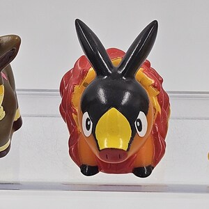 May include: A small, orange and black figurine of a Pokemon character with a yellow beak and black ears. The figurine is made of plastic and has a cartoonish style.