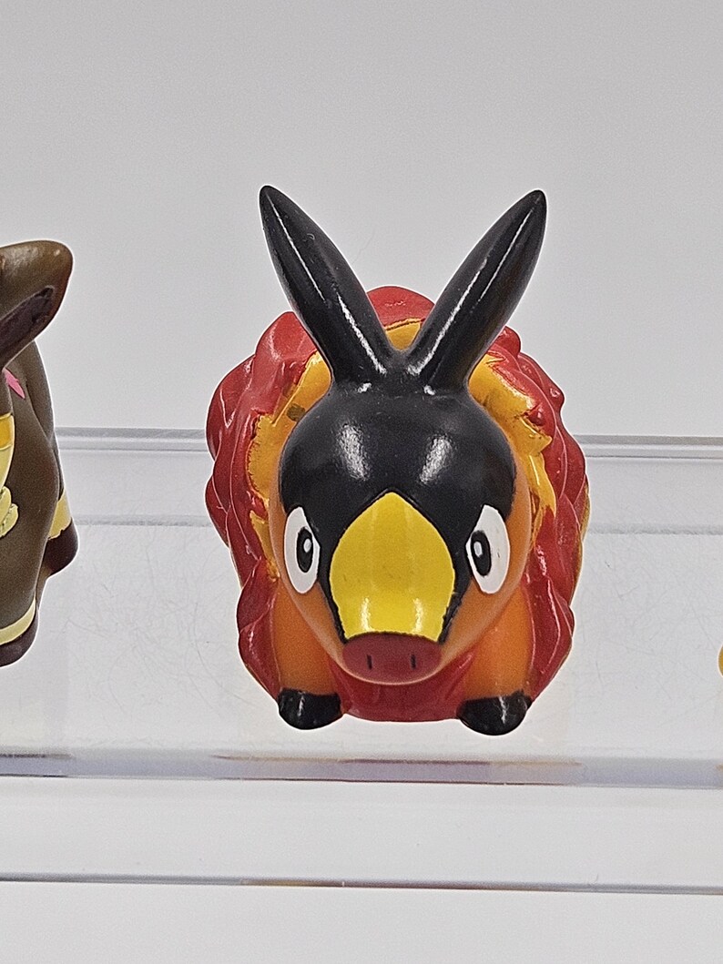 May include: A small, orange and black figurine of a Pokemon character with a yellow beak and black ears. The figurine is made of plastic and has a cartoonish style.