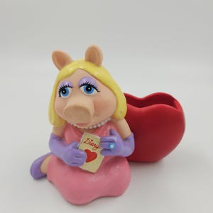 May include: A Miss Piggy figurine in a pink dress and purple gloves, holding a diary with a heart on the cover. A red heart-shaped container sits beside her. The figurine has blonde hair and blue eyes.