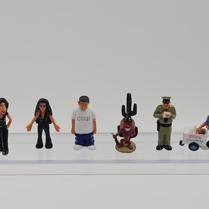 Może przedstawiać: Kolekcja miniaturowych figurek, w tym kobieta w czerni, mężczyzna w białej koszuli z nadrukiem "HOMIES", figurka diabła z kaktusem, policjant i sprzedawca lod&oacute;w z w&oacute;zkiem. Figurki znajdują się na przezroczystej p&oacute;łce.