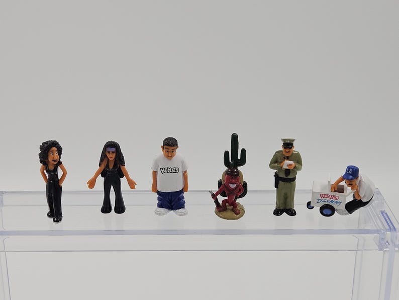 Może przedstawiać: Kolekcja miniaturowych figurek, w tym kobieta w czerni, mężczyzna w białej koszuli z nadrukiem "HOMIES", figurka diabła z kaktusem, policjant i sprzedawca lod&oacute;w z w&oacute;zkiem. Figurki znajdują się na przezroczystej p&oacute;łce.