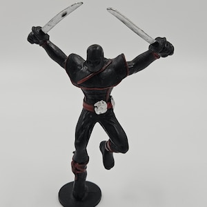 May include: A black action figure of a ninja-like character, poised in a dynamic pose with two silver swords held high. The figure has red accents on the shoulders, waist, and ankles. It stands on a black circular base.