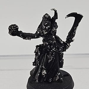 May include: A black miniature figurine standing on a circular base, featuring a skeletal figure wearing robes. The figure holds a skull in its outstretched right hand and a curved scythe in its left hand, with white highlights on the black surface.