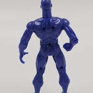 May include: A blue action figure of a humanoid robot, viewed from the back. The figure has a smooth, metallic appearance with articulated joints. The robot has a detailed design with a ribbed neck, and a segmented torso. The figure is standing on a white surface.