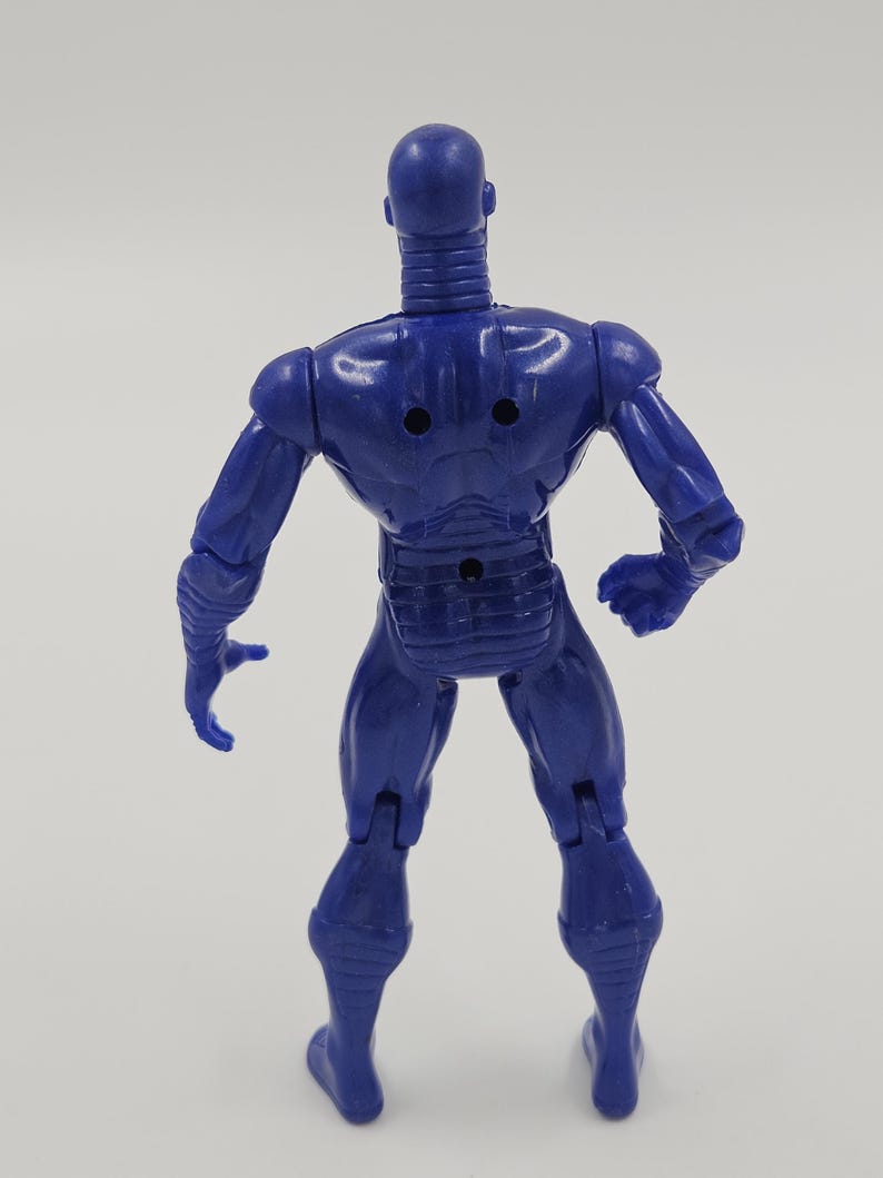 May include: A blue action figure of a humanoid robot, viewed from the back. The figure has a smooth, metallic appearance with articulated joints. The robot has a detailed design with a ribbed neck, and a segmented torso. The figure is standing on a white surface.