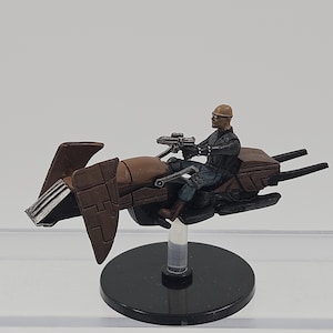 May include: A miniature figurine of a rider in a black jacket and tan helmet operating a brown and black speeder bike. The vehicle features angular side panels, a metallic front grill, and rear exhaust pipes, all mounted on a clear stand.