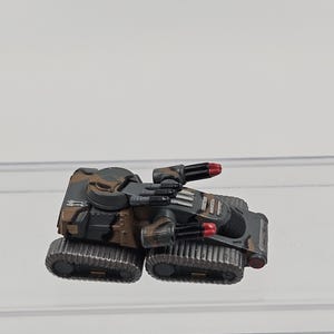 May include: A miniature tank model featuring a camouflage paint scheme in shades of brown, black, and gray. The vehicle is equipped with dual black cannons that have red tips, a rotating turret, and two sets of dark gray tank treads on each side.