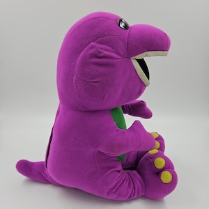 Peut inclure: Une peluche dinosaure violette avec un ventre vert, une large bouche ouverte montrant des dents blanches et de grands yeux noirs et blancs. La figurine a une petite queue, des bras courts et des pieds avec des accents circulaires jaunes sous les semelles.