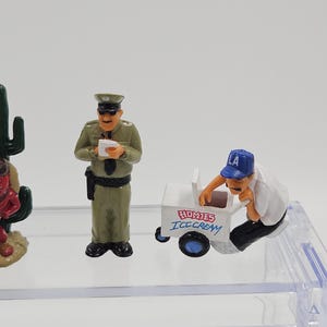 Może przedstawiać: Miniaturowe figurki policjanta, sprzedawcy lod&oacute;w z w&oacute;zkiem i kaktusa. Oficer nosi zielony mundur i trzyma papier. W&oacute;zek z lodami jest biały z niebieskimi kołami i napisem "HOMIES ICE CREAM".