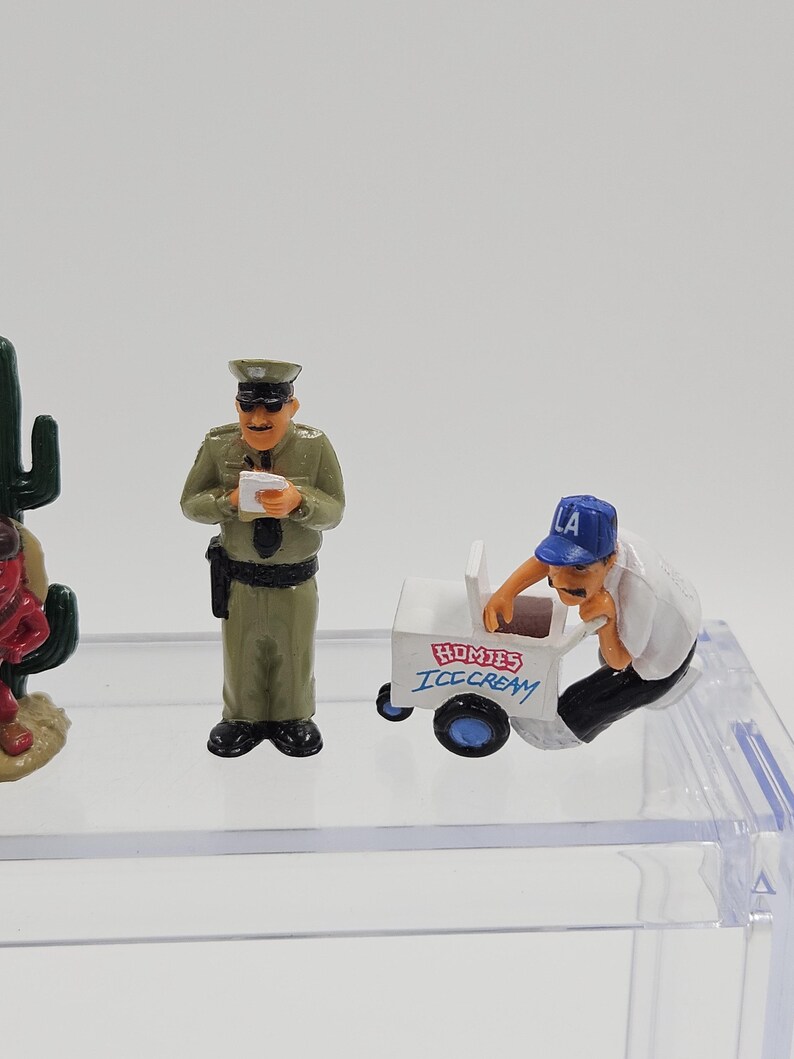 Może przedstawiać: Miniaturowe figurki policjanta, sprzedawcy lod&oacute;w z w&oacute;zkiem i kaktusa. Oficer nosi zielony mundur i trzyma papier. W&oacute;zek z lodami jest biały z niebieskimi kołami i napisem "HOMIES ICE CREAM".