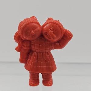 May include: A small, red plastic figurine of two children sharing a single body. The children are wearing matching outfits, including hats and dresses. The figurine has a vintage aesthetic.