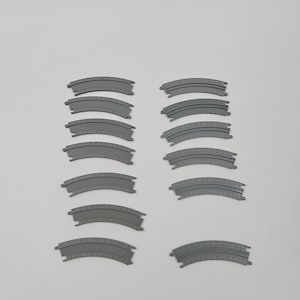May include: A collection of twelve grey plastic curved toy train track segments arranged in two vertical columns. Each individual track piece features a textured surface with raised parallel lines and interlocking connectors at both ends.