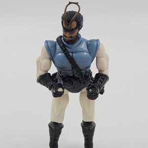 May include: A vintage action figure of a muscular character with a blue armored chest, black gloves, and boots. The figure has a dark beard, purple-tinted eyes, and a headpiece. The figure's legs are off-white.