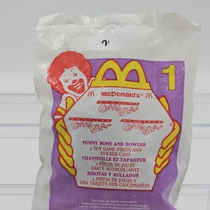 May include: A sealed McDonald's Crazy Bones toy package. The package features Ronald McDonald, the McDonald's logo, and the text "FUNNY BONE AND HOWLER". It includes two toy game pieces and a sticker card. The package is primarily white and purple.