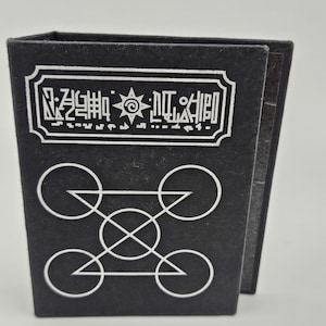 May include: Black, rectangular box with white geometric designs and text. The top of the box features a white rectangular label with stylized text and a sun-like symbol. Below, white circles and triangles form a complex pattern.