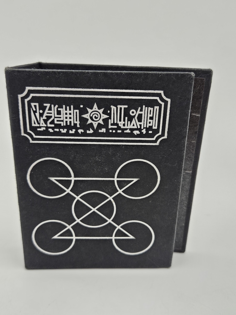 May include: Black, rectangular box with white geometric designs and text. The top of the box features a white rectangular label with stylized text and a sun-like symbol. Below, white circles and triangles form a complex pattern.