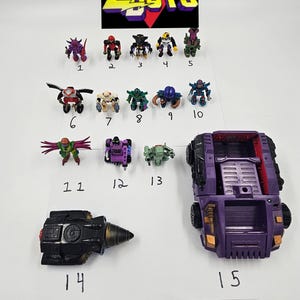 May include: A collection of 15 Micro Machines Zbots toys. The toys are arranged in rows on a white surface. The toys are mostly purple and black, with some red, green, and yellow accents. The toys are numbered 1 through 15.