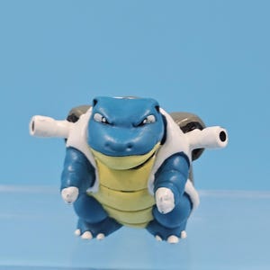 May include: A small, blue and yellow creature figurine with a turtle-like appearance, featuring two large cannons mounted on its back, white claws on its feet, and a stern facial expression with narrow eyes, standing against a solid blue background.