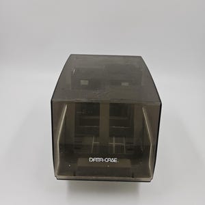 Puede incluir: Un contenedor de almacenamiento DATA-CASE translúcido de color gris oscuro. El contenedor tiene forma rectangular con una tapa con bisagras. El texto "DATA-CASE" está impreso en la parte frontal del contenedor.