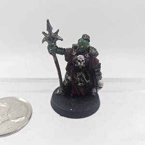 May include: A miniature fantasy figure of an orc, painted in green, red, and silver. The orc is holding a staff with a star-shaped top. The figure is on a black base and has a skull emblem on its chest. A coin is next to the figure.