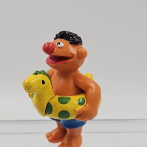 以下が含まれることがあります： オレンジ色の肌、黒い髪、赤い鼻をしたキャラクターのフィギュア。青いショートパンツを着用しています。キャラクターは、恐竜の形をした黄色と緑のインフレータブルリングを持っています。フィギュアはコレクターズトイです。
