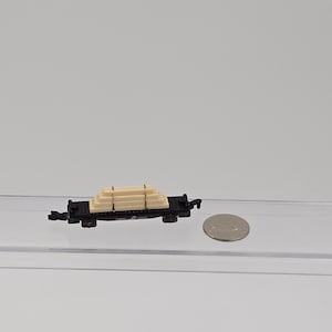 May include: A miniature black model train flatcar carrying a stack of light beige rectangular lumber pieces secured with thin black bands, positioned next to a silver-colored coin for scale on a clear surface.