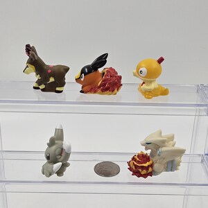 May include: Five small plastic figurines of Pokemon characters displayed on a two-tiered clear acrylic shelf. The top shelf features a brown deer-like Pokemon, a red and orange Pokemon with a black head, and a yellow bird-like Pokemon. The bottom shelf features a grey Pokemon with a long snout and a white Pokemon with a red and orange tail.