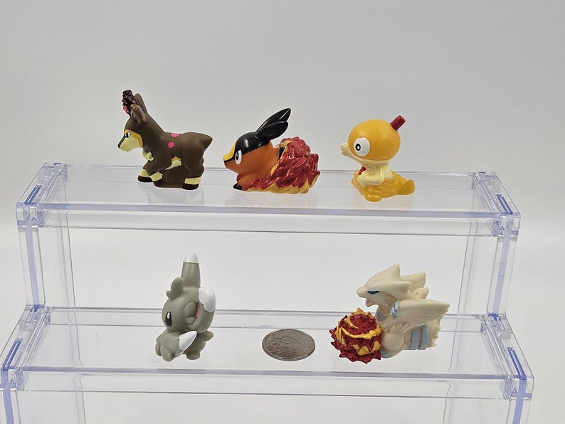 May include: Five small plastic figurines of Pokemon characters displayed on a two-tiered clear acrylic shelf. The top shelf features a brown deer-like Pokemon, a red and orange Pokemon with a black head, and a yellow bird-like Pokemon. The bottom shelf features a grey Pokemon with a long snout and a white Pokemon with a red and orange tail.