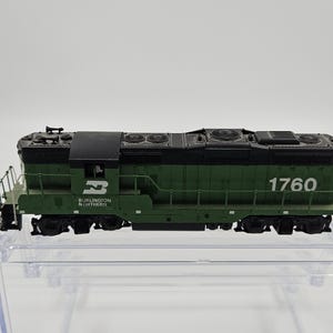Può includere: Una locomotiva di treno modello verde e nera con il numero 1760 in bianco. Le parole "Burlington Northern" sono stampate sul lato. Il modello di treno è su una superficie di plastica trasparente.
