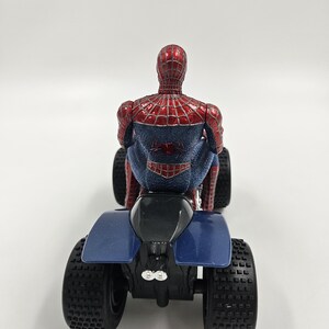 May include: A red and blue Spider-Man action figure riding a blue and black ATV. The figure is detailed with a web pattern and the Spider-Man logo on the back. The ATV has large, black, textured tires.