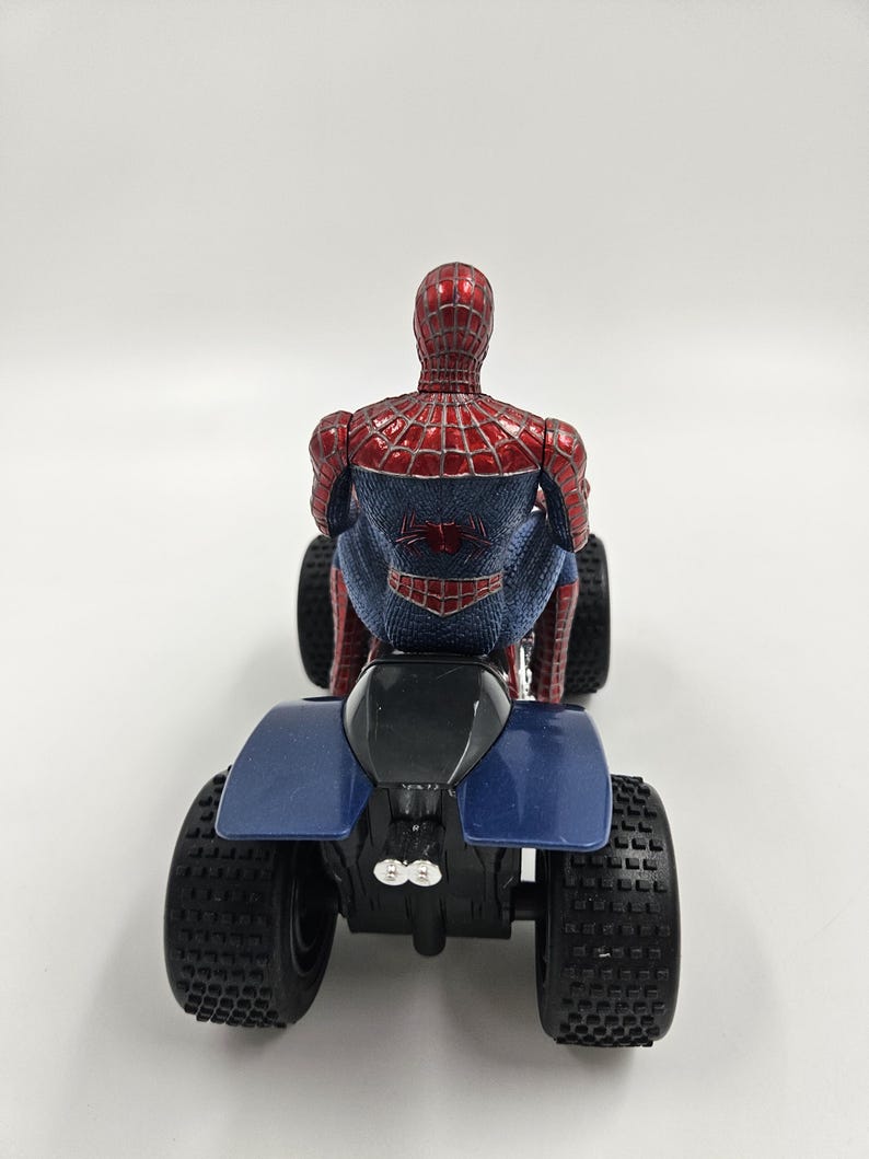May include: A red and blue Spider-Man action figure riding a blue and black ATV. The figure is detailed with a web pattern and the Spider-Man logo on the back. The ATV has large, black, textured tires.