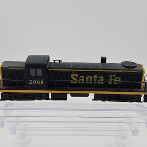 May include: A black and yellow Santa Fe model train. The train has the number 2099 on the side and the words "Santa Fe" in yellow lettering. The model train is sitting on a clear plastic display case.