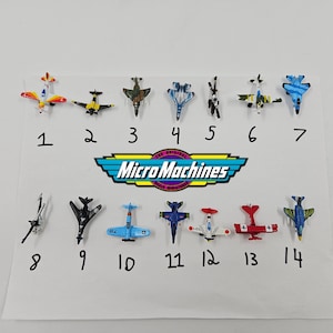 Puede incluir: Una colección de catorce aviones de juguete Micro Machines de varios colores y diseños, dispuestos sobre una superficie blanca. El logotipo de Micro Machines es visible. Cada avión está numerado del 1 al 14.