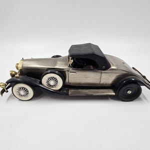 Puede incluir: Modelo de coche descapotable vintage de color plateado con guardabarros negros y capota negra. El coche tiene neumáticos de banda blanca y detalles dorados en la parte delantera. El modelo es una réplica detallada de un automóvil clásico.