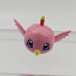 Puede incluir: Una figura de pájaro rosa de estilo caricatura con ojos azules, pico amarillo y pequeñas alas rosas. Una cresta roja y amarilla se encuentra en la parte superior de la cabeza del pájaro. El pájaro es un artículo decorativo caprichoso.