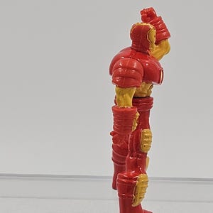 May include: A small plastic action figure of a character in a red and yellow armored suit. The figure is shown from a side profile, standing upright with one arm raised near the head, featuring molded details on the chest, arms, and legs.