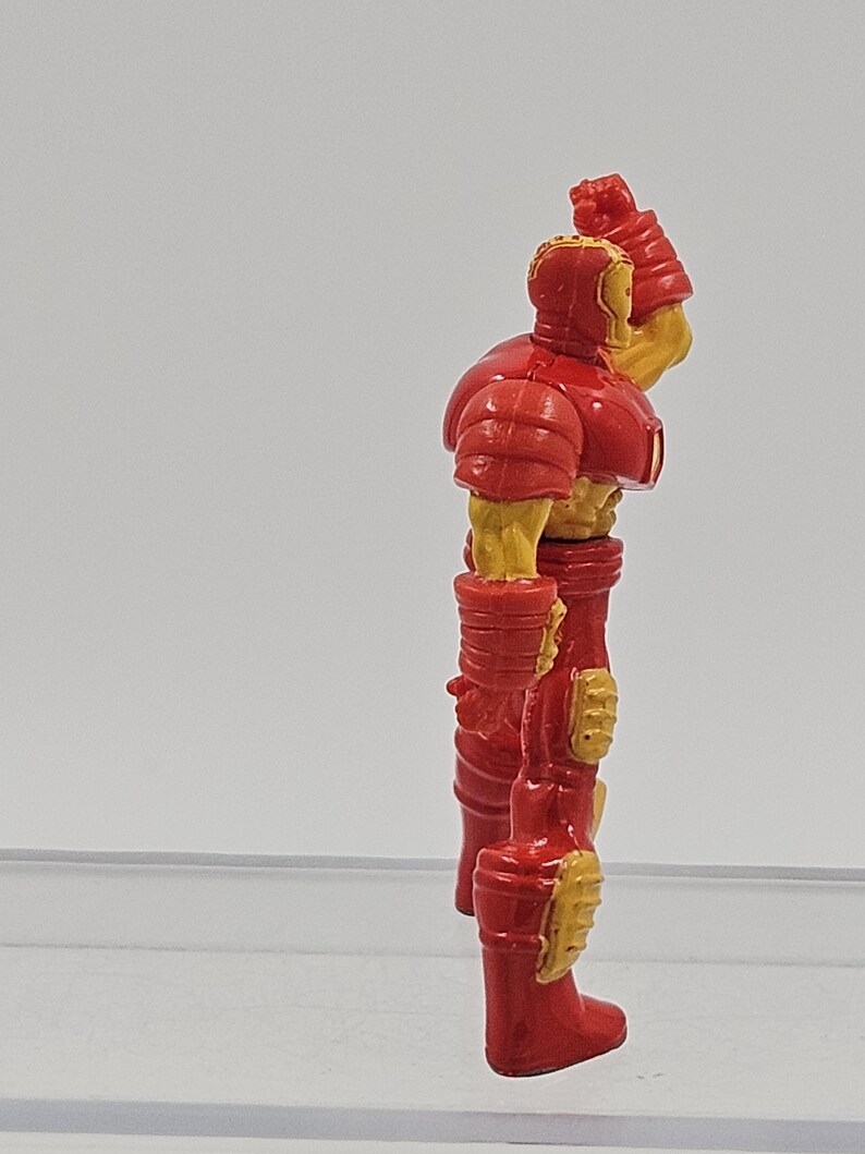 May include: A small plastic action figure of a character in a red and yellow armored suit. The figure is shown from a side profile, standing upright with one arm raised near the head, featuring molded details on the chest, arms, and legs.