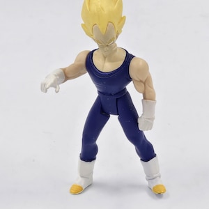May include: A yellow-haired action figure of a character wearing a blue jumpsuit and white boots. The figure is posed in a fighting stance with its arms outstretched.