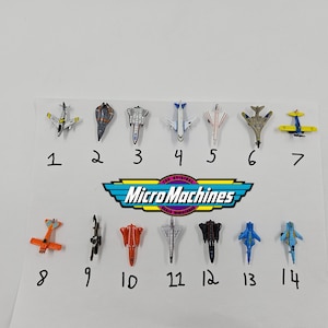 Puede incluir: Una colección de catorce aviones de juguete Micro Machines en varios colores, incluyendo naranja, azul, plateado y negro. El logotipo de Micro Machines es visible, junto con números que identifican cada avión.