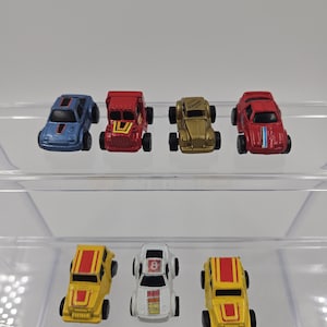 Puede incluir: Una colección de ocho coches de juguete en miniatura de varios colores, incluyendo rojo, azul, amarillo, blanco y dorado. Los coches están dispuestos en dos filas sobre una superficie de plástico transparente. Algunos de los coches tienen rayas de carreras y números.